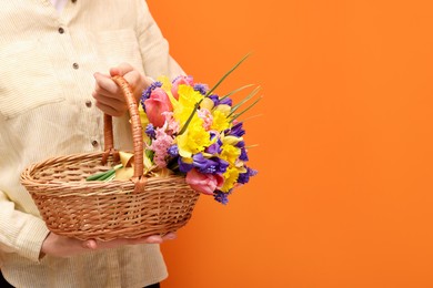Photo of Woman holding wicker basket with beautiful flowers on orange background, closeup. Space for text