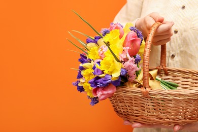 Photo of Woman holding wicker basket with beautiful flowers on orange background, closeup. Space for text