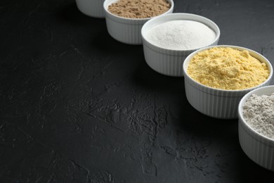 Photo of Different types of flour in bowls on black textured table, closeup. Space for text