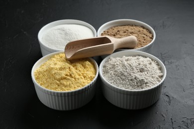 Photo of Different types of flour in bowls and scoop on black textured table, closeup