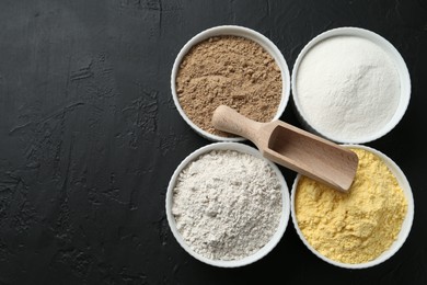 Photo of Different types of flour in bowls and scoop on black textured table, top view. Space for text