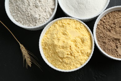 Photo of Different types of flour in bowls and spike on black table, flat lay