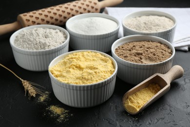 Photo of Different types of flour in bowls, scoop and rolling pin on black textured table, closeup
