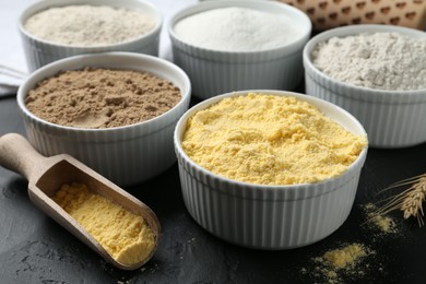 Photo of Different types of flour in bowls and scoop on black textured table, closeup