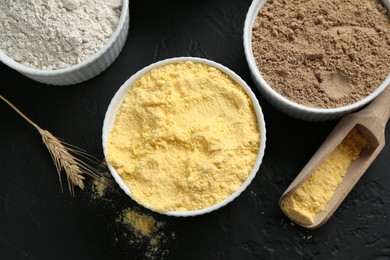 Photo of Different types of flour in bowls, scoop and spike on black textured table, flat lay