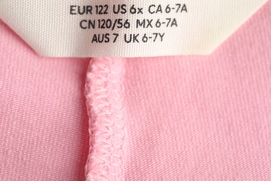 Photo of Clothing label with size on pink garment as background, closeup