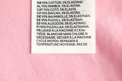 Photo of Clothing label with care advice and size on pink garment as background, top view