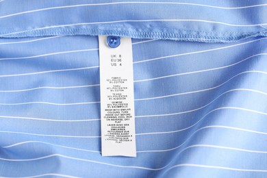 Photo of Clothing label with material content and size on light blue garment as background, above view