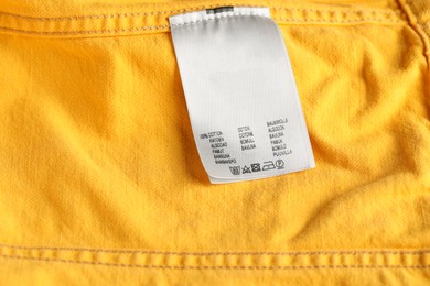 Photo of Clothing label with care symbols and material content on yellow garment as background, top view