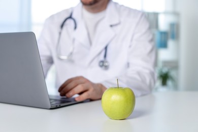 Photo of Nutritionist working with laptop at table in clinic, focus on green apple