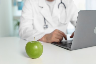 Photo of Nutritionist working with laptop at table in clinic, focus on green apple