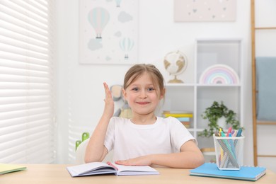 Photo of Happy little girl raising hand while sitting at table indoors