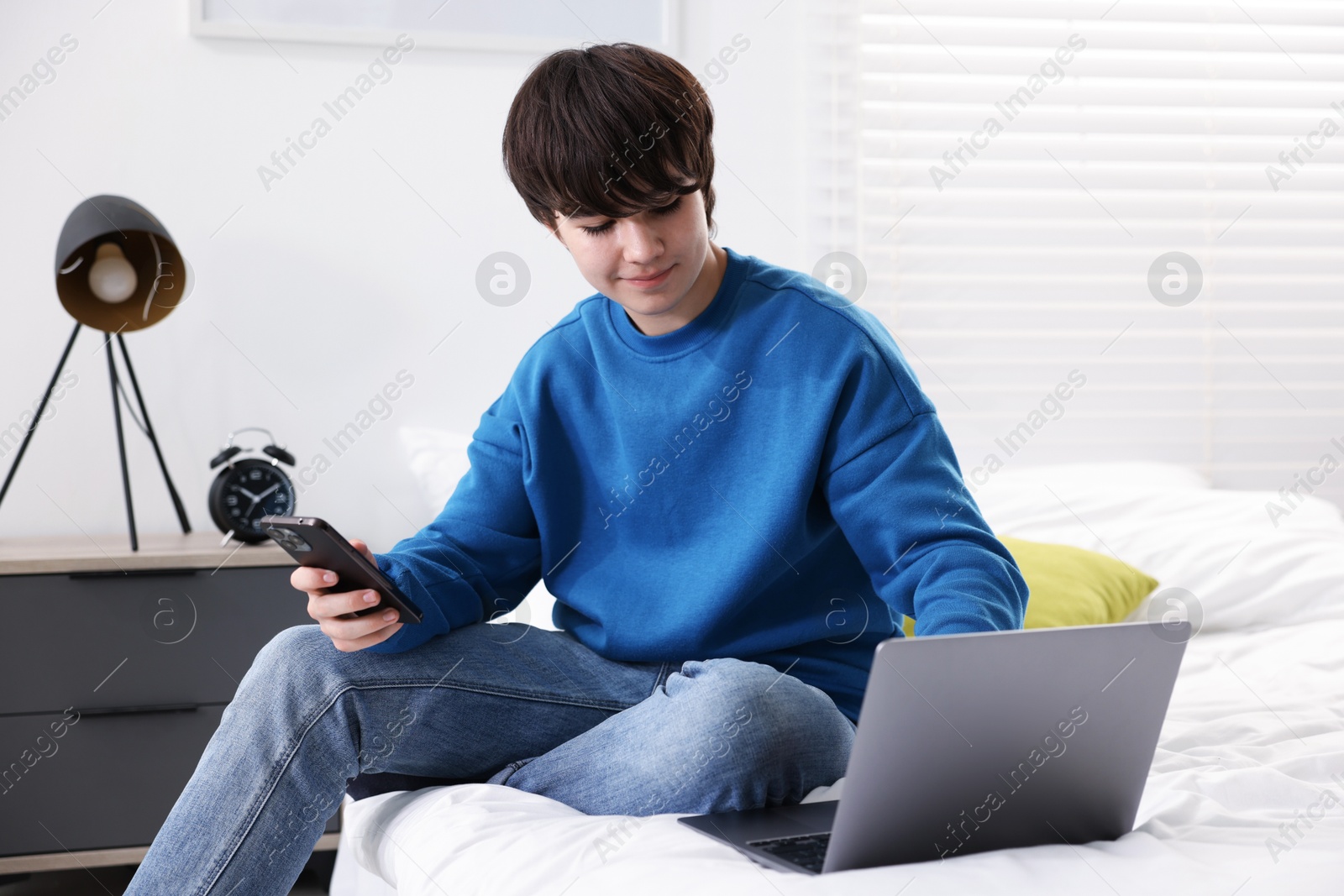 Teenage boy with smartphone and laptop on bed at home Photo of Teenage boy with smartphone and laptop on bed at home