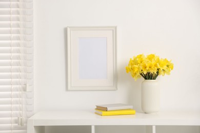Photo of Beautiful daffodils in vase and books on white cabinet at home