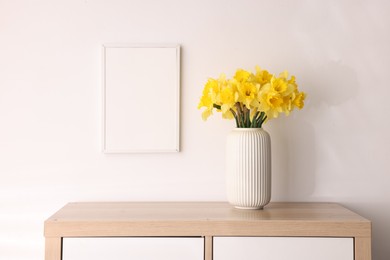 Photo of Beautiful daffodils in vase and picture frame on cabinet at home