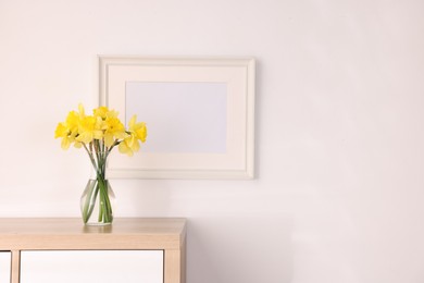 Photo of Beautiful daffodils in vase and picture frame on cabinet at home, space for text
