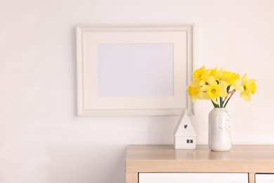 Photo of Beautiful daffodils in vase, stylish home decor and picture frame on cabinet in room