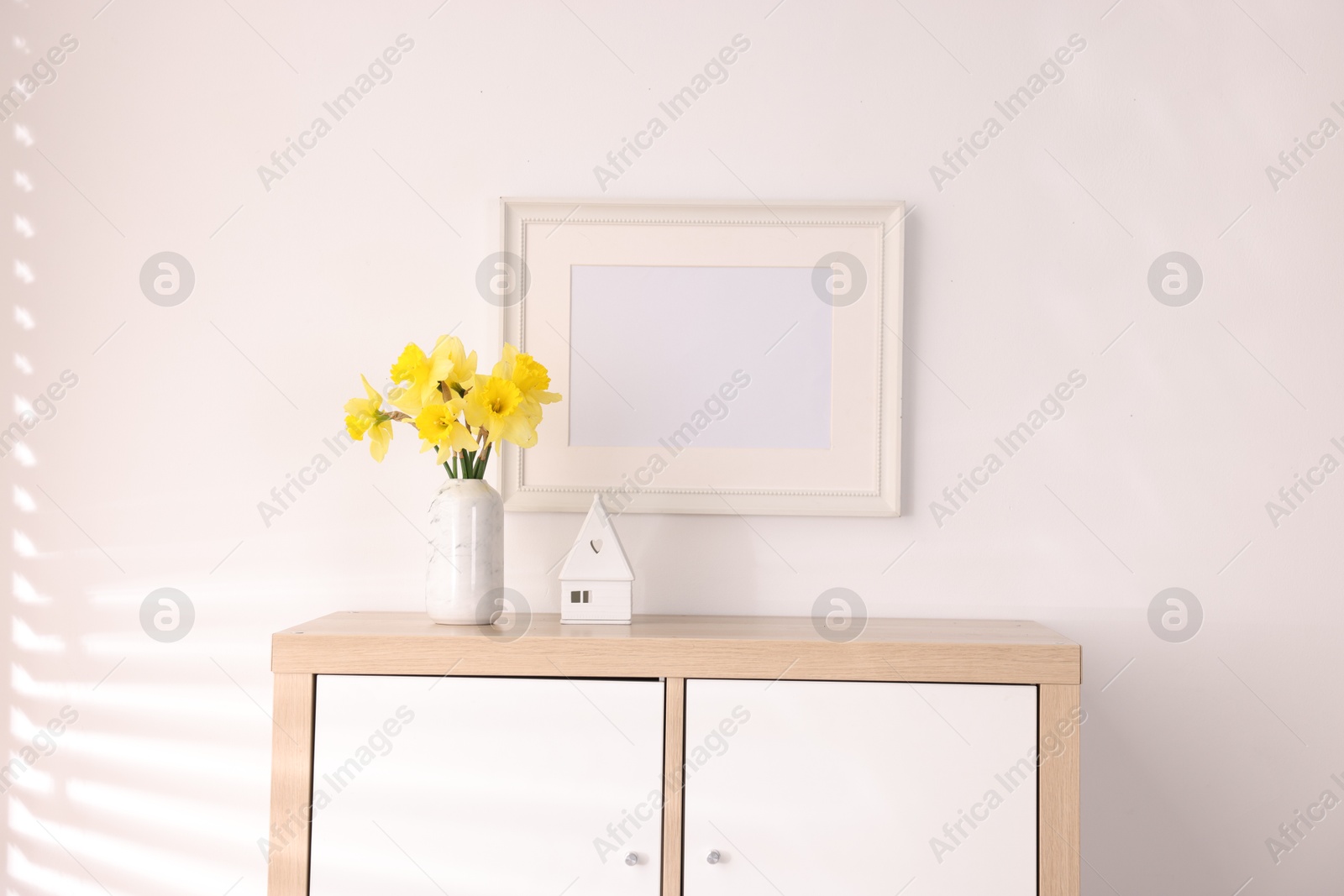 Beautiful daffodils in vase, stylish home decor and picture frame on cabinet in room Photo of Beautiful daffodils in vase, stylish home decor and picture frame on cabinet in room