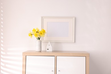 Photo of Beautiful daffodils in vase, stylish home decor and picture frame on cabinet in room