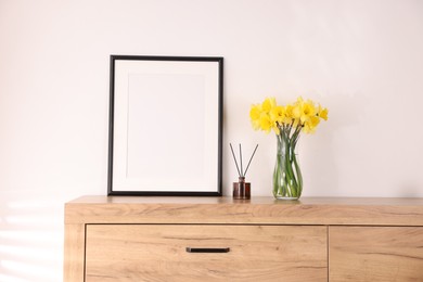 Photo of Beautiful daffodils in vase, reed diffuser and picture frame on cabinet at home