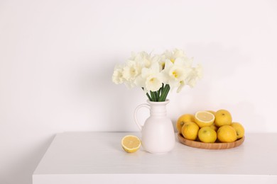 Photo of Beautiful daffodils in vase and lemons on white wooden table. Space for text