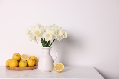Photo of Beautiful daffodils in vase and lemons on white wooden table. Space for text