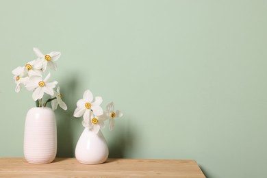 Photo of Beautiful daffodils in vases on wooden table near light green wall. Space for text