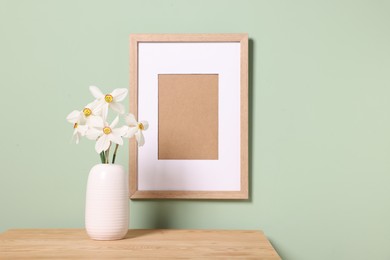 Photo of Beautiful daffodils in vase and picture frame on wooden table near light green wall