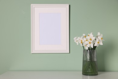 Photo of Beautiful daffodils in vase and picture frame on white table near light green wall