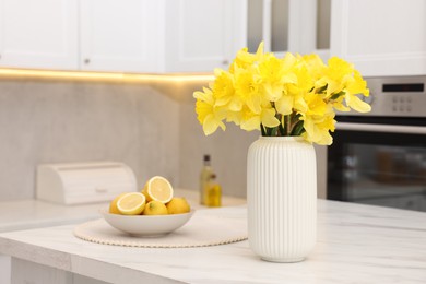 Photo of Beautiful daffodils in vase and lemons on white marble table in kitchen