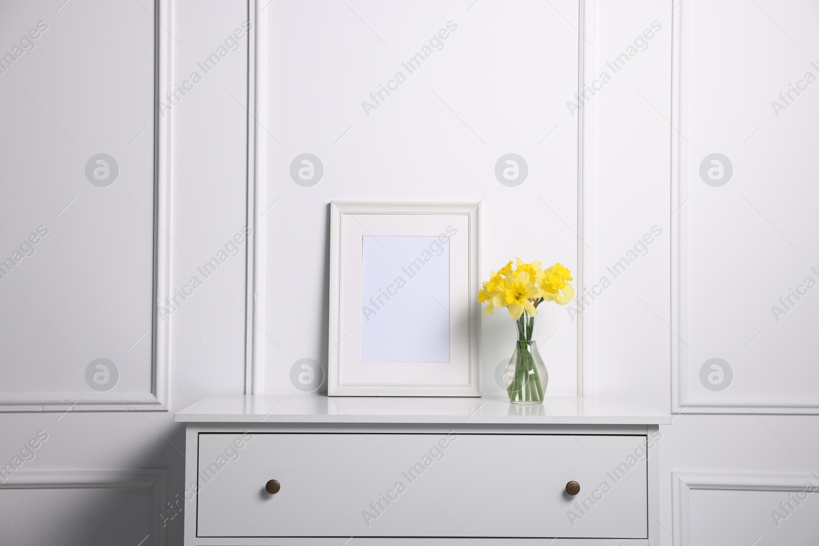 Beautiful daffodils in vase and picture frame on cabinet near white wall at home Photo of Beautiful daffodils in vase and picture frame on cabinet near white wall at home