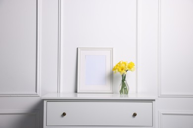 Photo of Beautiful daffodils in vase and picture frame on cabinet near white wall at home