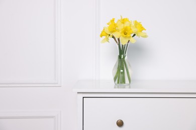 Photo of Beautiful daffodils in vase on cabinet near white wall at home. Space for text