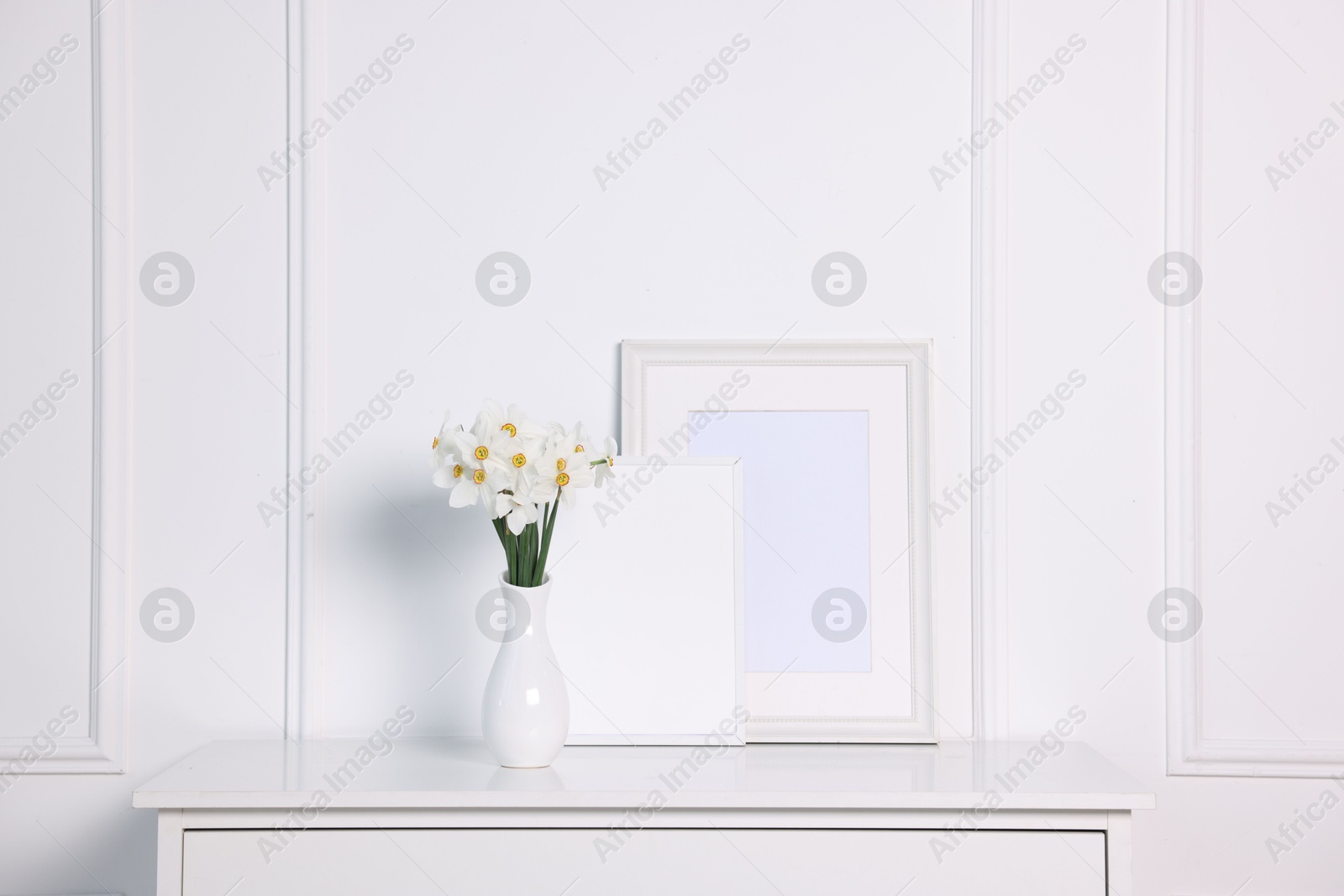 Beautiful daffodils in vase and picture frames on cabinet near white wall at home Photo of Beautiful daffodils in vase and picture frames on cabinet near white wall at home