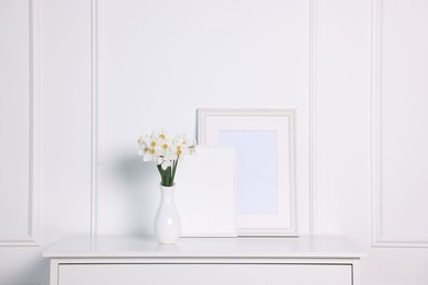 Photo of Beautiful daffodils in vase and picture frames on cabinet near white wall at home
