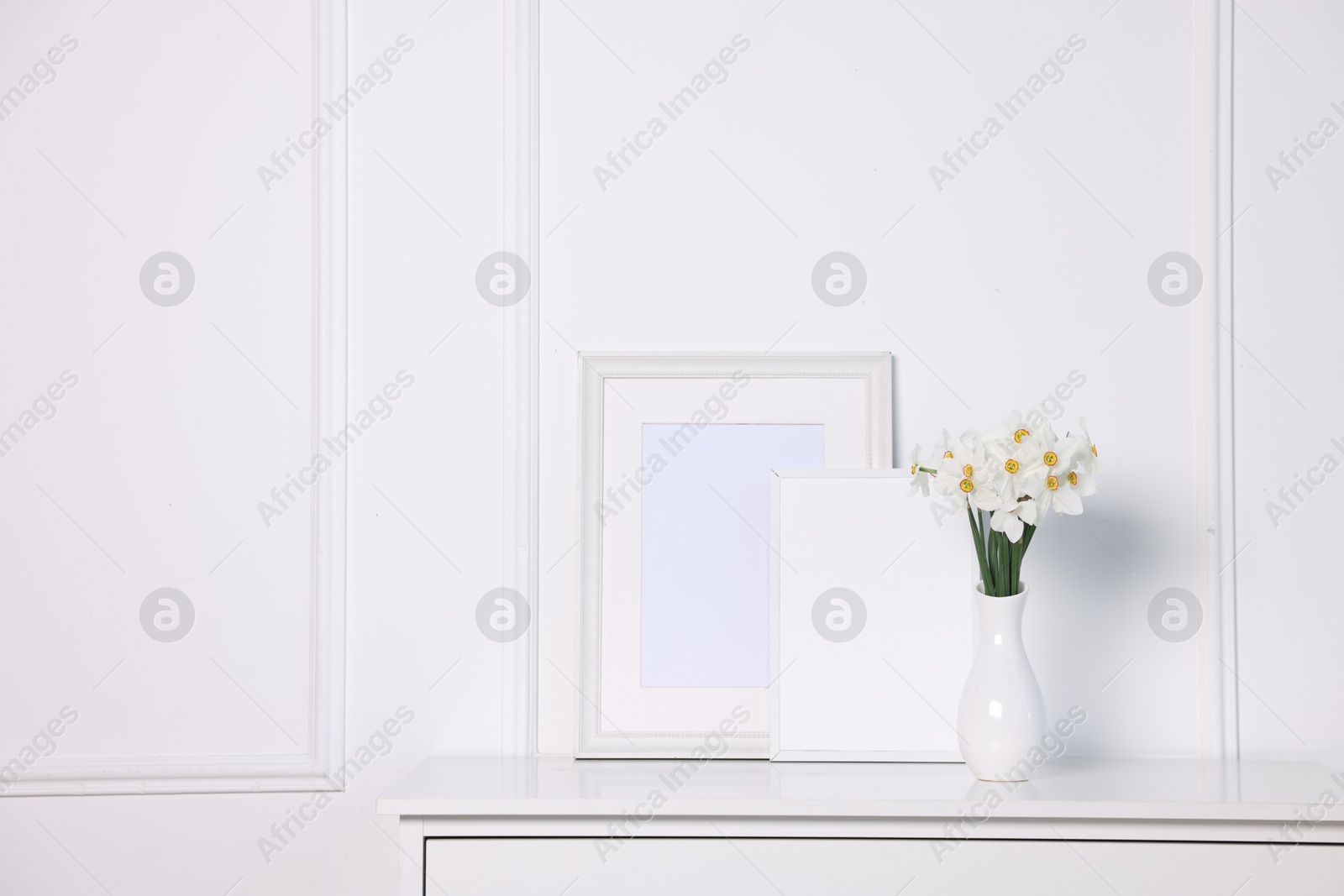 Beautiful daffodils in vase and picture frames on cabinet near white wall at home. Space for text Photo of Beautiful daffodils in vase and picture frames on cabinet near white wall at home. Space for text