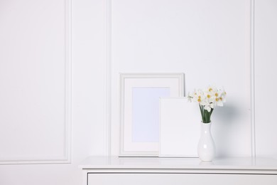 Photo of Beautiful daffodils in vase and picture frames on cabinet near white wall at home. Space for text