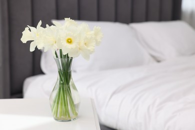 Photo of Beautiful daffodils in vase on white table near bed at home. Space for text
