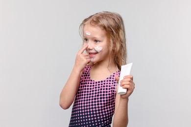 Photo of Smiling little girl with tube of sun protection product applying cream onto her nose against grey background