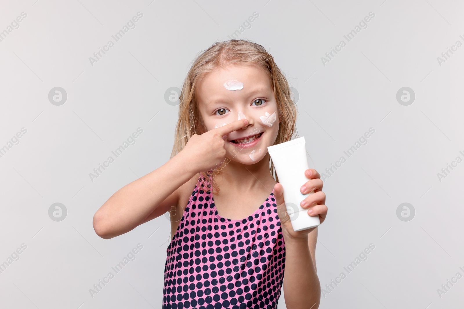 Smiling little girl with tube of sun protection product applying cream onto her nose against grey background Photo of Smiling little girl with tube of sun protection product applying cream onto her nose against grey background