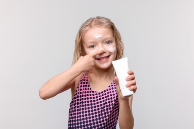 Photo of Smiling little girl with tube of sun protection product applying cream onto her nose against grey background