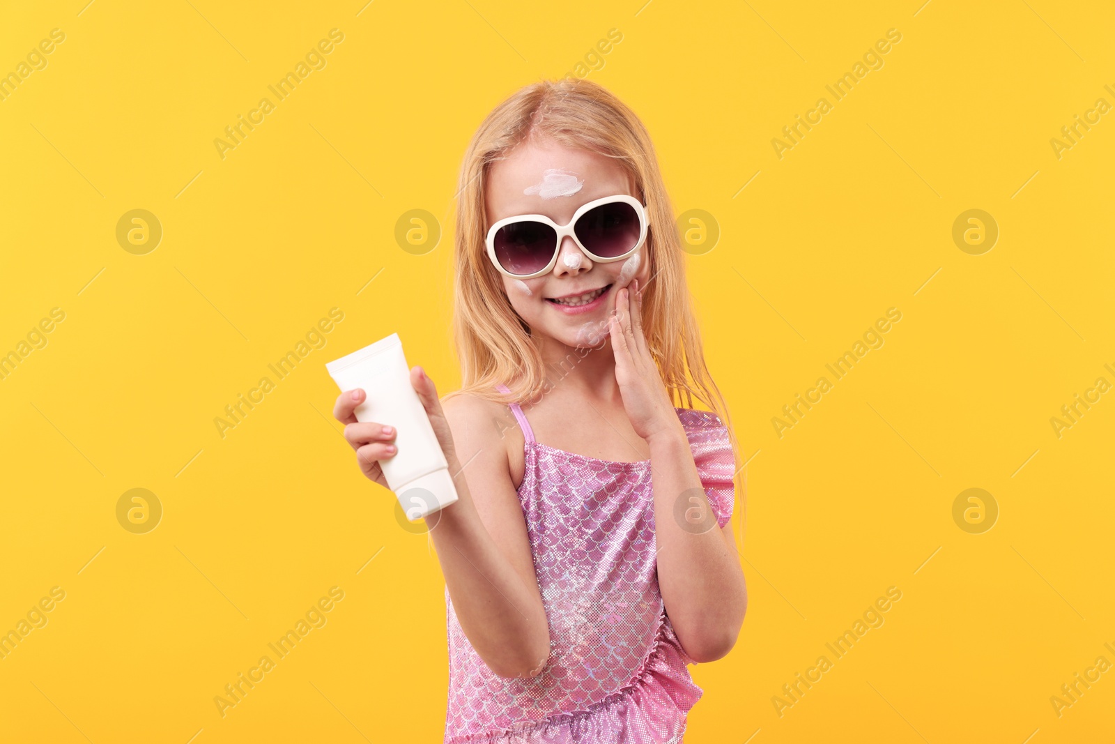 Smiling little girl with tube of sun protection cream against yellow background Photo of Smiling little girl with tube of sun protection cream against yellow background