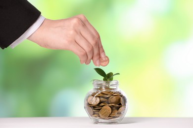 Photo of Investment concept. Woman putting coin into glass jar at light table against blurred green background, closeup