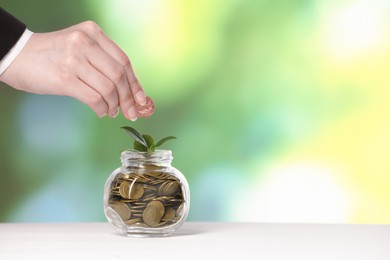 Photo of Investment concept. Woman putting coin into glass jar at light table against blurred green background, closeup. Space for text