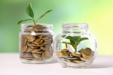 Photo of Investment concept. Glass jars with coins and sprouts on light table against blurred green background, selective focus