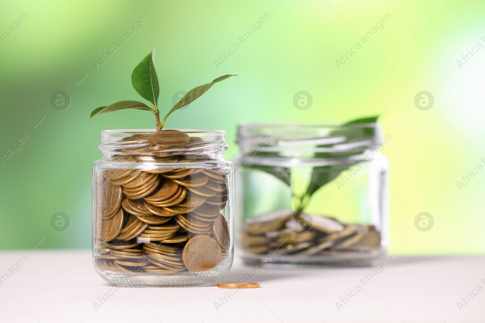 Investment concept. Glass jars with coins and sprouts on light table against blurred green background, selective focus Photo of Investment concept. Glass jars with coins and sprouts on light table against blurred green background, selective focus
