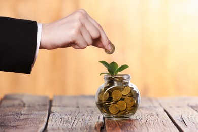 Photo of Investment concept. Woman putting coin into glass jar at wooden table against blurred orange background, closeup