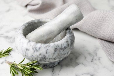 Photo of Mortar with rosemary, pestle and cloth on white marble table, closeup