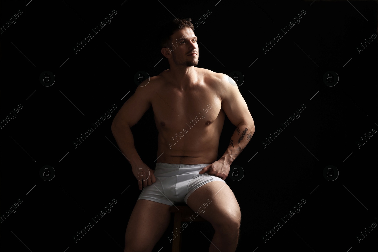 Young man in stylish white underwear on chair against black background Photo of Young man in stylish white underwear on chair against black background