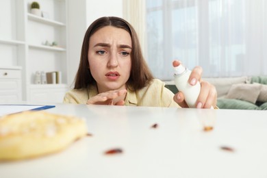 Photo of Emotional young woman spraying insecticide onto cockroaches at white table indoors. Pest problem